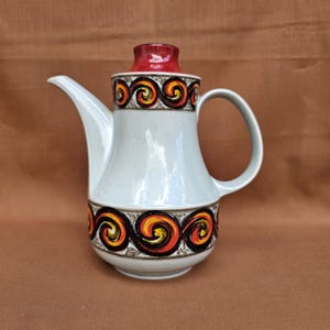 Puede incluir: Una cafetera de cerámica vintage con cuerpo gris claro, tapa roja y asa. La cafetera presenta una banda decorativa de patrones de remolinos naranja, amarillo y negro en la parte superior e inferior. El pico es largo y curvado.