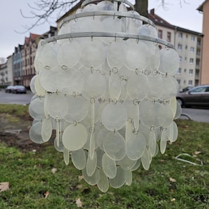 Könnte beinhalten: Eine dekorative Außenleuchte in Birnenform. Die Leuchte besteht aus zahlreichen durchscheinenden, kreisförmigen Scheiben, die an einem Metallrahmen aufgehängt sind. Der obere Teil hat ein zylindrisches beiges Element. Im Hintergrund ist ein Wohngebiet zu sehen.