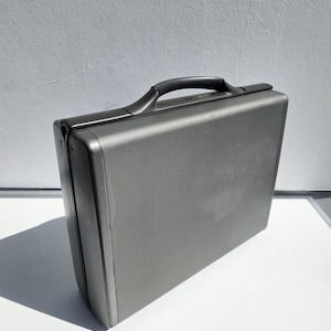 May include: A gray, rectangular briefcase with a textured surface and a black handle. The briefcase is standing upright on a white surface, with a gray wall in the background. The briefcase appears to be made of metal.
