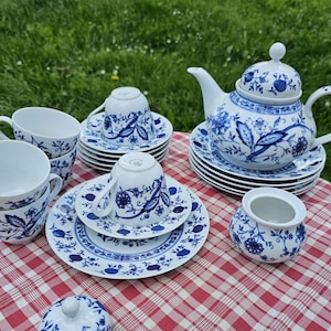 Può includere: Un servizio da tè in porcellana bianca con un motivo floreale blu. Il set comprende una teiera, tazze, piattini e una zuccheriera. Il servizio da tè è disposto su una tovaglia a scacchi rossi e bianchi.