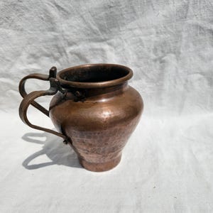 Puede incluir: Una jarra de cobre martillado con dos asas. La jarra tiene un cuerpo bulboso y un interior oscuro. El cobre tiene un tono marrón rojizo cálido y una superficie texturizada. Las asas están unidas al cuerpo con remaches.