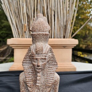 May include: A detailed, brown stone bust of an Egyptian pharaoh, featuring a traditional headdress and intricate facial details. The sculpture is set against a backdrop of dried reeds and a terracotta-colored planter.