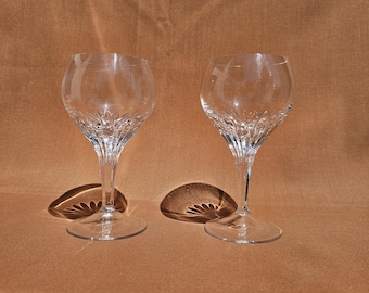 May include: Two clear crystal wine glasses with a detailed, textured design on the lower bowl and stem. The glasses are positioned on a brown surface, casting shadows. The stemware has a classic, elegant shape.