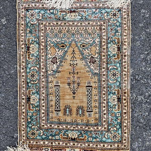 May include: A small, rectangular rug with a detailed, ornate design. The rug features a central tan field with a stylized arch and floral motifs, framed by a teal and cream border. White fringe edges the rug.