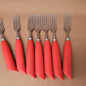 Könnte beinhalten: Ein Set aus sieben Vintage-Gabeln mit Edelstahlzinken und leuchtend roten Griffen. Die Gabeln sind auf einer braunen Oberfläche angeordnet und zeigen ihr klassisches Design und ihre lebendige Farbe.