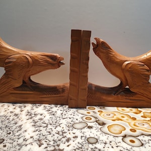 May include: A pair of wooden bookends carved in the shape of birds with their wings spread. The bookends are a light brown color and have a rustic finish.