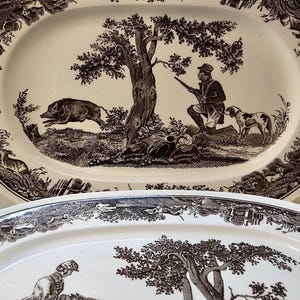 Op de afbeelding: Twee ovale serveerschalen met een sepia-getinte jachtscène. De gedetailleerde illustratie toont een jager, honden en een wild zwijn in een bosrijke omgeving. De schalen hebben een decoratieve rand.