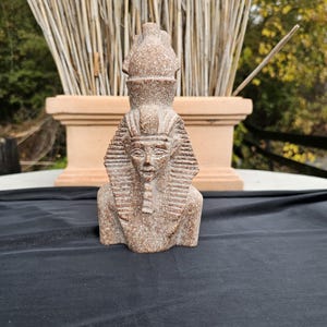 May include: A detailed, brown stone sculpture of an Egyptian pharaoh bust. The bust features a detailed headdress, a serene facial expression, and a traditional collar. The sculpture is set against a backdrop of natural elements, including a terracotta planter and tall, dried reeds.