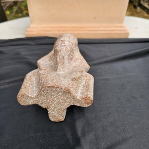May include: A detailed, stone sculpture of a pharaoh's head, featuring a headdress and intricate facial details. The sculpture is a light brown color and rests on a black surface. A terracotta-colored planter is in the background.
