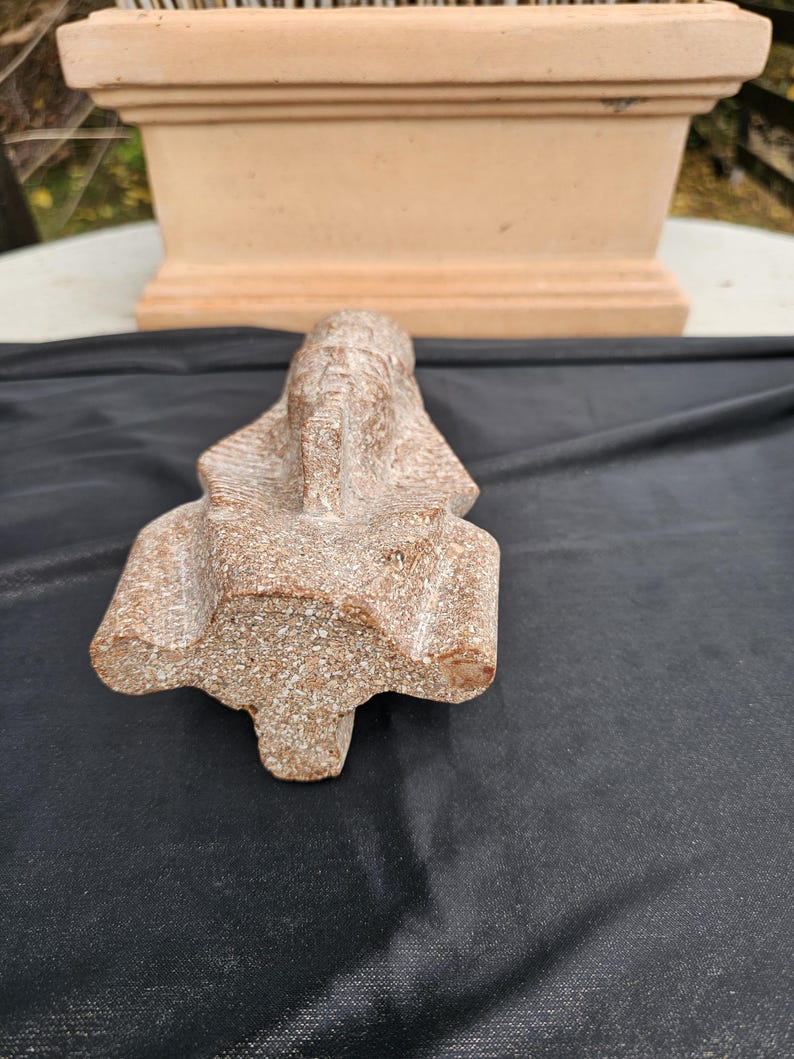 May include: A detailed, stone sculpture of a pharaoh's head, featuring a headdress and intricate facial details. The sculpture is a light brown color and rests on a black surface. A terracotta-colored planter is in the background.