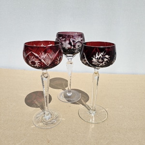 May include: Three stemmed glasses featuring deep red bowls with intricate cut-glass star and floral patterns. Each glass has a clear, faceted stem and a circular base with a radial sunburst design, showcasing craftsmanship in colored crystal glass.