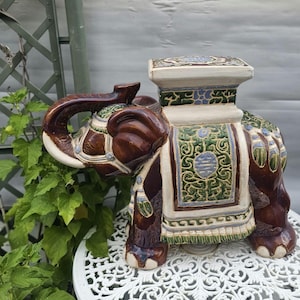 May include: A ceramic elephant garden stool featuring a glossy dark brown glaze, a cream-colored rectangular top, and a decorative saddle blanket with intricate green, blue, and white patterns. The elephant has a raised trunk and detailed harness.
