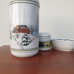 Könnte beinhalten: Ein Set aus drei weißen Keramikbehältern mit einem Wintermotiv. Der größte Behälter hat einen grünen Deckel und zeigt eine rote Scheune, Bäume und eine verschneite Landschaft. Die beiden anderen Behälter sind kleiner und haben ein grün-weißes Design.
