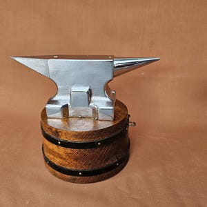 Op de afbeelding: Een miniatuur zilveren aambeeld met een puntige hoorn, gemonteerd op een houten vatvormige basis. De basis is gemaakt van donker hout met zwarte banden en kleine metalen studs. Het aambeeld is een symbool van smeden.