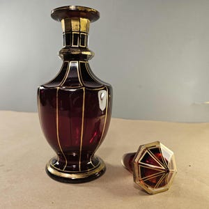 May include: A ruby red glass decanter with gold accents and a matching stopper. The decanter has a bulbous body and a flared base. The stopper is an octagonal shape with gold trim. The decanter is likely vintage.