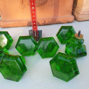 Può includere: Una collezione di otto pomelli per mobili in vetro verde. Ogni pomello ha una forma esagonale e una vite color ottone. I pomelli sono disposti su una superficie bianca, con un righello rosso sullo sfondo. I pomelli hanno un diametro di circa 3,8 cm.