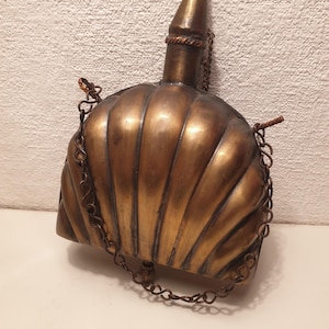 Op de afbeelding: Een vintage messing fles met een schelpvormig lichaam en een puntige bovenkant. Het heeft een ketting die aan de boven- en onderkant is bevestigd.