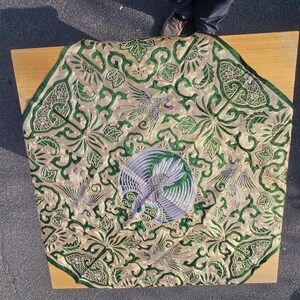 Pode incluir: Têxtil dourado ornamentado com bordados verdes intrincados. O design apresenta pássaros estilizados e motivos florais. O têxtil é colocado sobre uma superfície de madeira castanha clara.