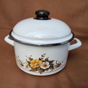 May include: White enamel pot with lid and side handles. The pot features a floral design with yellow and white flowers. The lid has a black knob and a silver rim. A vintage kitchenware item.