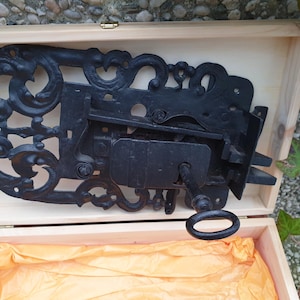 Könnte beinhalten: Ein verziertes schwarzes Schloss aus Metall mit einem Schlüsselloch und einem dekorativen Schlüssel in einer Holzkiste mit orangefarbenem Seidenpapier.
