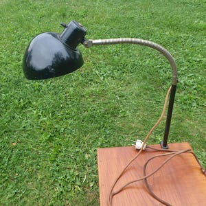 May include: A black desk lamp with a flexible arm and a black shade. The lamp is plugged into an outlet and the cord is coiled on a wooden surface.