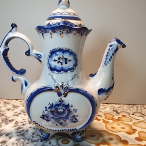 May include: A white porcelain teapot with blue and gold floral designs. The teapot has a round body with a heart-shaped design on the front. The teapot has a lid with a finial and a spout on the side. The teapot is sitting on a white surface with brown and white patterns.