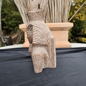 May include: A carved stone sculpture of a figure with a tall headdress and detailed facial features. The sculpture is a light brown color and has a textured surface. The figure is standing upright.