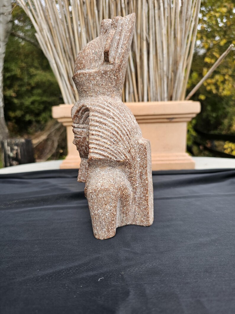 May include: A carved stone sculpture of a figure with a tall headdress and detailed facial features. The sculpture is a light brown color and has a textured surface. The figure is standing upright.