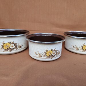 Op de afbeelding: Drie vintage emaille kommen met een crèmekleurige basis en een bloemenontwerp met gele en witte bloemen. De kommen hebben bruine interieurs en zilveren randen, perfect voor keukeninrichting.