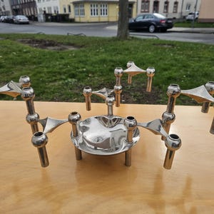May include: A silver-colored, multi-arm candle holder with a central dish. The candle holder has multiple arms extending outwards, each with a cylindrical candle cup. The design is modern and geometric, with a polished finish.