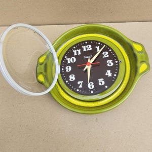 May include: A round green ceramic wall clock with a dark brown face and white numerals. The clock features a hinged clear plastic cover that is open to the left. The hands are gold-toned with a red second hand, and the word QUARZ is printed in white.