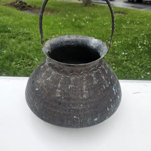 May include: A dark, weathered metal bucket with a rounded body and a swinging handle. The surface features a hammered texture, visible oxidation with green patina spots, and an engraved decorative pattern around the upper rim area.