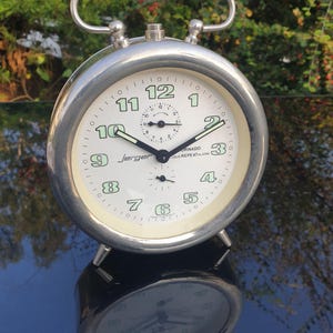 May include: A silver alarm clock with a white face and black hands. The clock has a large, round face with black numbers and a small, round alarm clock face. The clock is labeled "Jerger Tornado Loud Repeat Alarm".