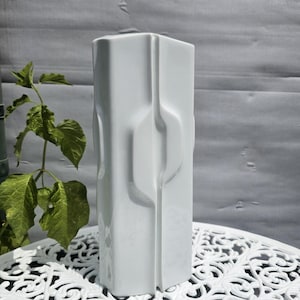 Könnte beinhalten: Eine hohe, weiße Keramikvase mit glatter, glänzender Oberfläche. Die Vase zeichnet sich durch ein geometrisches, kantiges Design mit vertikalen, vertieften Kanälen und erhabenen Paneelen aus, die ein strukturiertes, facettiertes Aussehen erzeugen.