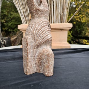 May include: A reddish-brown stone sculpture of a seated figure with a tall headdress. The figure has carved details on the chest and head. The sculpture is on a black surface.