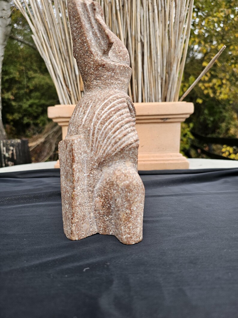 May include: A reddish-brown stone sculpture of a seated figure with a tall headdress. The figure has carved details on the chest and head. The sculpture is on a black surface.
