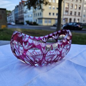 Könnte beinhalten: Eine dekorative, oval geformte Kristallschale in tiefem Magenta. Die Schale weist komplizierte Blumen- und geometrische Muster auf, und ein kleiner, runder Aufkleber ist an der Seite sichtbar. Die Schale liegt auf einer weißen Oberfläche.