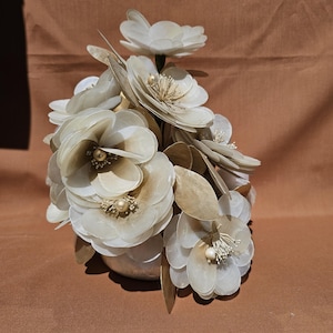 Könnte beinhalten: Ein Strauß künstlicher Blumen in einer silbernen Vase. Die Blumen sind weiß und cremefarben mit goldenen Akzenten und perlenartigen Zentren. Die Anordnung umfasst mehrere große, offene Blüten und Blätter.