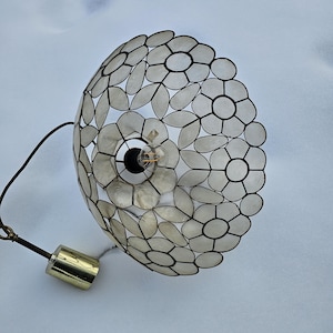 Può includere: Una lampada a sospensione decorativa con un design floreale. Il paralume è composto da elementi a forma di petalo bianco sporco, delineati in nero, disposti in un motivo circolare. Un elemento cilindrico dorato pende dall'apparecchio.