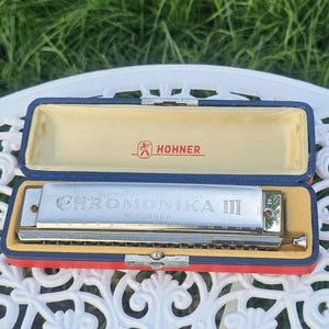 Peut inclure: Un harmonica Hohner Chromonika III argenté reposant dans un étui rectangulaire ouvert bleu et rouge avec une doublure intérieure crème. L'harmonica présente du texte gravé sur sa surface métallique et le logo Hohner est imprimé en rouge à l'intérieur du couvercle.