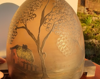 May include: A decorative, egg-shaped object with a landscape design. The artwork features a tree and a small house with a thatched roof, painted in shades of brown, gold, and black. The object is displayed on a wooden stand.