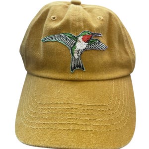 Op de afbeelding: Een mosterdgele baseballpet met een geborduurde kolibrie op het voorpaneel. De vogel is afgebeeld met groene vleugels, een rode keel en een witte borst. De pet heeft een gebogen klep met zichtbare stiksels en een knoop aan de bovenkant.