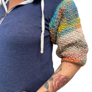 May include: A navy blue hooded sweatshirt with a crocheted sleeve featuring a rainbow of colors. The sleeve is crocheted in a striped pattern with shades of blue, green, yellow, orange, and brown.