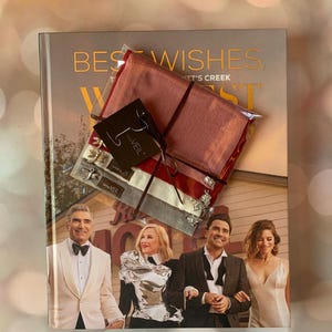 May include: A book titled "Best Wishes, The Schitt's Creek" with a red, white, and gray scarf wrapped around it. The scarf has a small tag that says "The Veil". The book cover features a photo of the four main characters from the show.