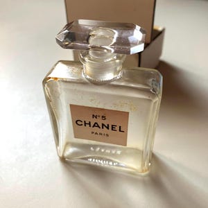 May include: A clear glass perfume bottle with a rectangular shape and a faceted stopper. The label reads "N&deg;5 CHANEL PARIS". The bottle is partially filled with a light-colored liquid. A cardboard box is in the background.
