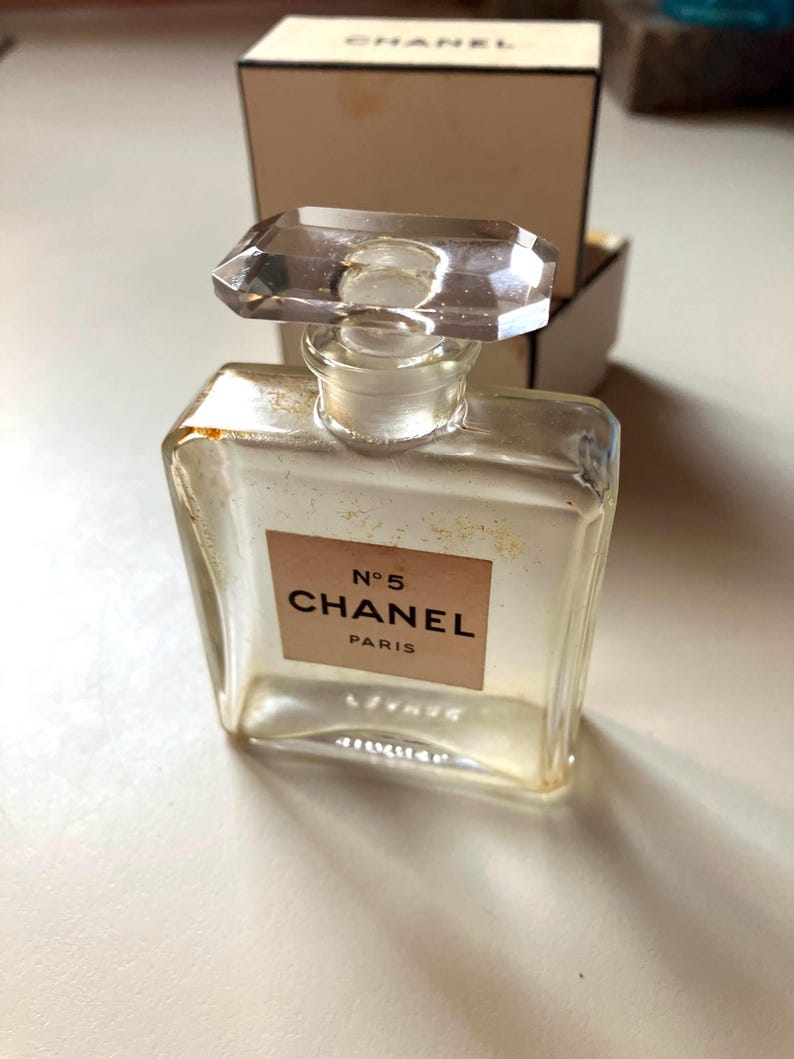 May include: A clear glass perfume bottle with a rectangular shape and a faceted stopper. The label reads "N&deg;5 CHANEL PARIS". The bottle is partially filled with a light-colored liquid. A cardboard box is in the background.