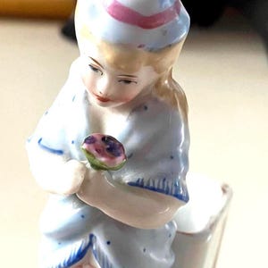 May include: A porcelain figurine of a person wearing a light blue hat with a pink stripe and a matching light blue garment with blue speckles and fringe details. The figure holds a small purple and green flower in their hands, looking down.