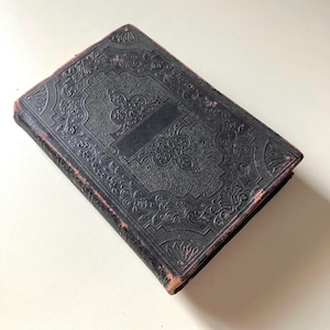 May include: A closed, antique book with a black, embossed cover featuring ornate floral and scroll designs. The edges of the book show wear and a reddish-brown color. The book is rectangular and appears to be leather-bound.