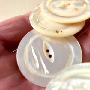 May include: A set of four white mother-of-pearl buttons with two holes each. The buttons have a smooth, shiny surface and a slightly iridescent sheen. One button has a carved design of two lines.
