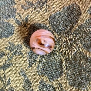 May include: A spherical glass marble featuring a translucent light pink base color with swirling streaks of dark brown and opaque white throughout the surface, creating a marbled effect within the smooth, polished, and reflective glass material.